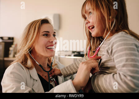 Smiling young pediatrician playing with a girl patient at her clinic. Friendly doctor and young girl examining each other with stethoscope. Stock Photo
