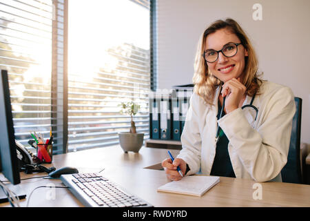 Smiling female doctor sitting her office desk with a notepad. Female doctor in white coat looking to camera in an office. Stock Photo