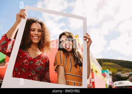 Two smiling woman standing together and holding empty frame outdoors. Beautiful female friends holding a blank picture frame looking at camera, smilin Stock Photo