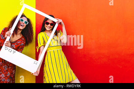 Portrait of two women holding a blank photo frame in hand and smiling. Girls wearing sunglasses standing against red and yellow colored wall. Stock Photo