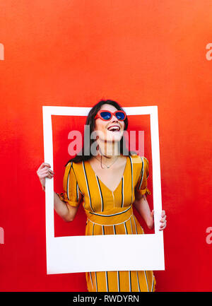 Stylish woman standing against the red wall and holding a large photo frame. Beautiful girl wearing sunglasses looking through blank picture frame. Stock Photo