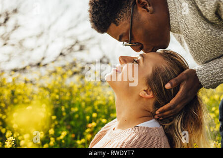 Close up of young man kissing forehead of woman. Romantic interracial couple outdoors in meadow. Stock Photo