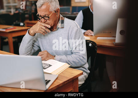 Elderly gentleman reading a book sitting in classroom with a laptop in front. Senior man learning in a university classroom. Stock Photo