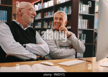 Two senior men sitting in a library and learning with books and a computer on the table. Senior learners discussing sitting in classroom. Stock Photo