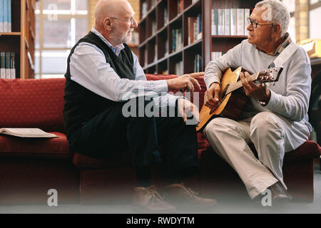 Two senior men sitting on couch and playing guitar together. Senior friends relaxing and playing music on guitar. Stock Photo
