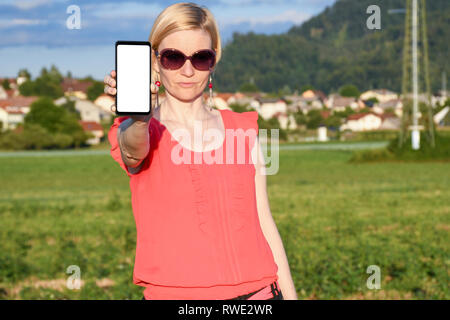 Beautiful woman wearing sun glasses while holding in hand a smartphone with a white screen on a countryside on a hot sunny summer day Stock Photo