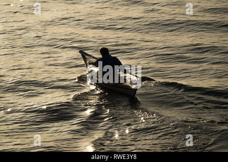 A solitary man paddling a kayak at sunset in silhouette on calm water with copy space water full frame