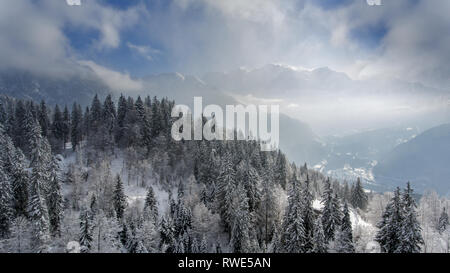 Snow covered winter trees in the foreground frame a perfect winter scene as a snowy alpine mountain tops peak through the clouds and mist. Stock Photo