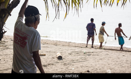 Old Fisherman watching younger generation pull net to beach from sea - Antique, Philippines Stock Photo