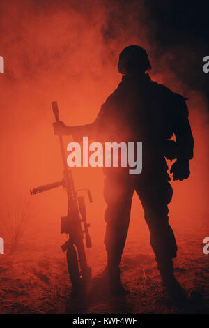 Backlit silhouette of Army sniper with large caliber rifle standing in the fire and smoke. Stock Photo