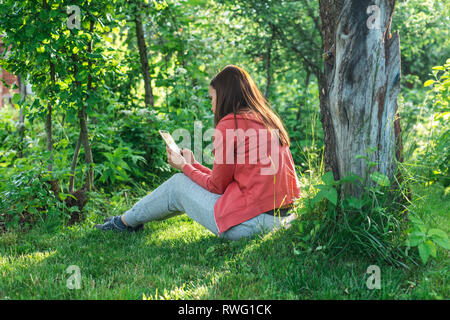 Girl in a sporty pink jacket and gray pants sits on a green spring lawn near a tree and reads on the phone Stock Photo