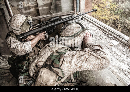 Sniper team shooting enemy targets with sniper rifle from inside a ruined urban building. Stock Photo