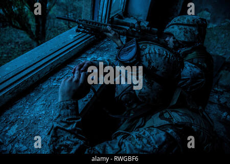 Sniper team with large caliber sniper rifle hiding in a ruined urban building at night. Stock Photo