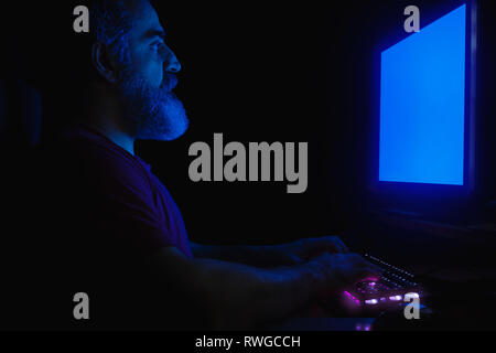 Bearded man sitting in front of computer monitor, hands on a keyboard, working, illuminated with blue light from monitor Stock Photo