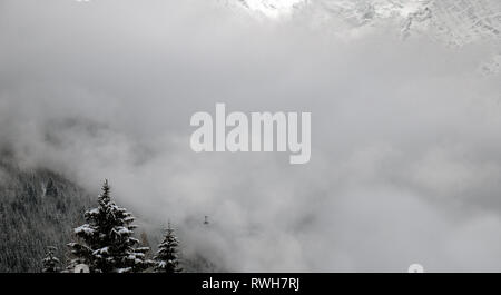 A cable car rises up the mountain in the french alpine town of Chamonix. The forests and mountains are covered in fresh snow and mist. Stock Photo