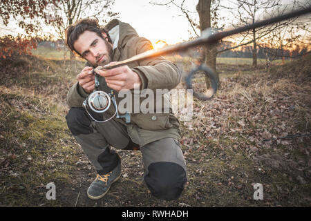 Young man kneeling outdoors in a thick jacket is checking a fishing rod Stock Photo