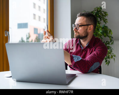 Home and communication concept - man in glasses sitting at home office and working on a laptop and using smartphone Stock Photo