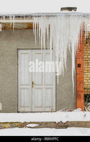 Huge icicles hang from the roof of an abandoned house with wooden door. Stock Photo