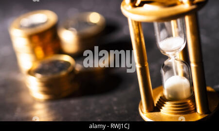 Sand trickles through an hourglass with coins in the background, symbolizing the time factor when investing money Stock Photo