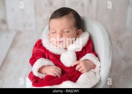 Portrait of one week old newborn baby girl sleeping in a chair and wearing a red and white Mrs. Claus dress. Stock Photo