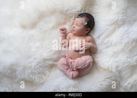 Sleeping, week old newborn baby girl wearing light pink, knitted pants and holding a tiny, heart shaped pillow. Shot in the studio on a white sheepski Stock Photo