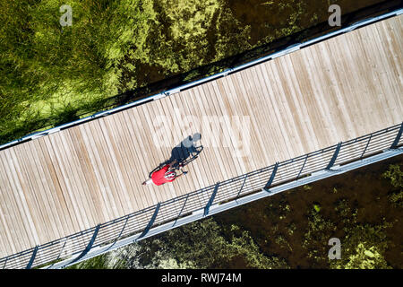 Aerial view looking straight down on a female cyclist on a bridge across a swampy pond with shadow of cyclist, East of Calgary; Alberta, Canada Stock Photo