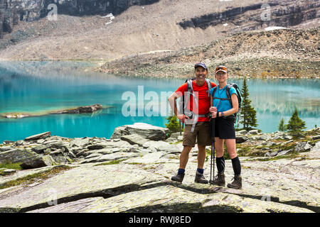 Female and male hiker standing in a large rocky area with colourful alpine lake and mountain cliffs in the background; British Columbia, Canada Stock Photo