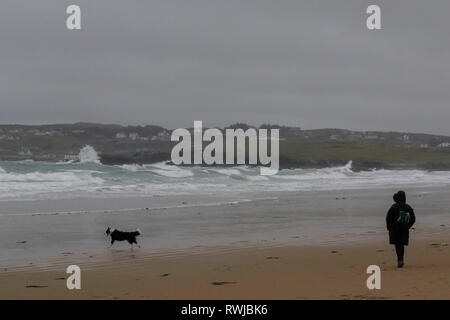 County Donegal, Ireland. 06 March 2019. Ireland weather - after heavy overnight rain, it was a day of strong winds and driving rain across County Donegal. Woman and dog on Dunfanaghy Beach as waves crash into the rocks towards Portnablagh. Woman with dog playing on beach with white waves in Ireland. Credit: David Hunter/Alamy Live News. Stock Photo