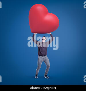 Young fit man in casual clothes holding up huge red Valentine heart on blue background. Stock Photo