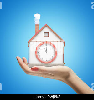 Female hand holding white house with a clock on blue background Stock Photo