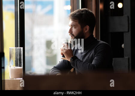 handsome pensive bearded guy near the window in a cafe. attractive young stylish man sitting in a cafe and looking out the window. Business, freelance Stock Photo