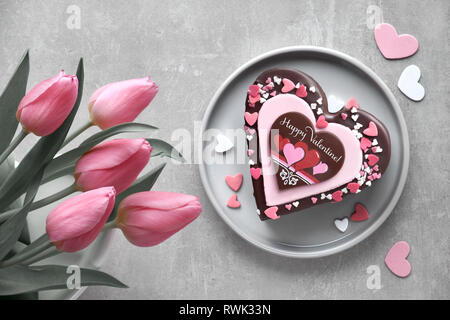 Valentine heart cake with chocolate, sugar decorations and text 'Happy Valeitine' and a bunch of pink tulips. Top view on light background. Stock Photo