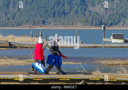 Middle aged couple in winter clothing walking with life jackets and kayak paddles along a pier in Grant Narrows Regional Park, B. C., Canada. Stock Photo