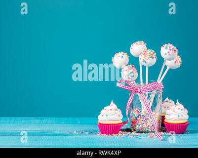Vanilla cake pops with colorful sprinkles and cupcakes on a blue background. Stock Photo