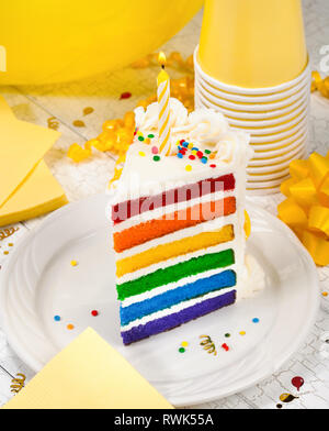 Slice of colourful birthday cake with lit birthday candle and yellow party decorations in the background Stock Photo