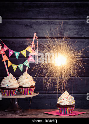 Cupcakes with Happy Birthday Banner, lit sparkler and colorful sprinkles on a dark background Stock Photo