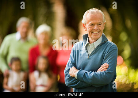 Senior man smiles proudly and stands with his arms crossed as he poses for a portrait with his family in the background. Stock Photo