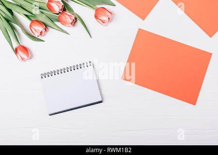 Flat lay floral arrangement of empty notebook next to blank red sheet of paper and bouquet of beautiful fresh tulips on white wooden background, top v Stock Photo