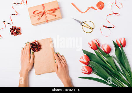 Female's hands decorating box with red bow near scissors, ribbons and bouquet of tulip flowers on white wooden table, top view. Festive flat lay arran Stock Photo