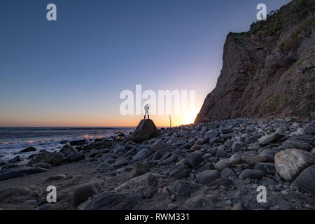 Beachside view of a man posed on a rock near a tall cliff as the sun approaches the horizon, Dana Point, California Stock Photo