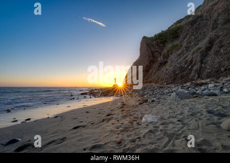 Beachside view of a man posed on a rock near a tall cliff as the sun approaches the horizon, Dana Point, California Stock Photo