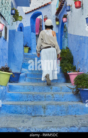 Back view of lady in skirt and hat going up on blue stairs near old houses in Marrakesh, Morocco Stock Photo