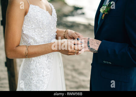 Bride putting on groom's wedding ring on lakeside, mid section Stock Photo