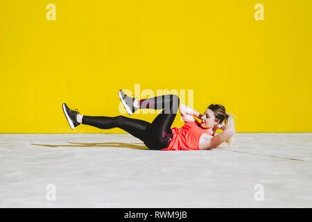 Young lady in sportswear doing sit ups exercises on floor near yellow wall Stock Photo