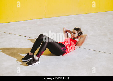 Young lady in sportswear doing sit ups exercises on floor near yellow wall Stock Photo