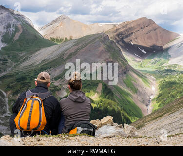 Male and female hiker sitting on a rock ridge overlooking a valley and mountain range in Kananaskis Country; Alberta, Canada Stock Photo