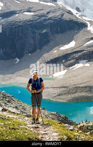 Female hiker on mountain pathway with a colourful alpine lake and mountain cliffs with snow in the background; British Columbia, Canada Stock Photo