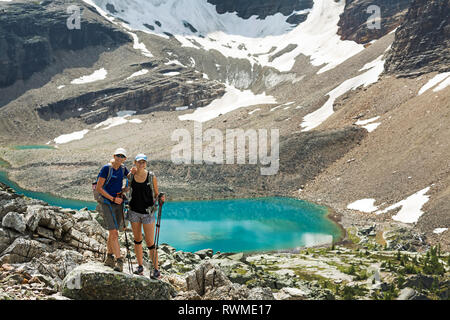 Two female hikers standing in a large rocky area with a colourful alpine lake and mountain cliffs with snow in the background Stock Photo