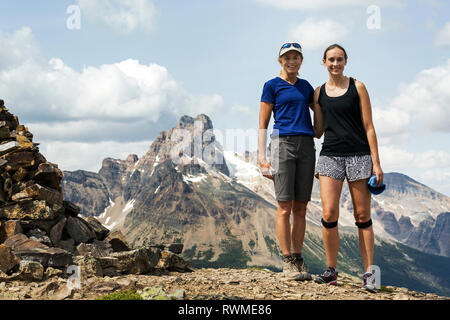 Two female hikers standing on rocky mountain top with mountain, blue sky and clouds in the background; British Columbia, Canada Stock Photo