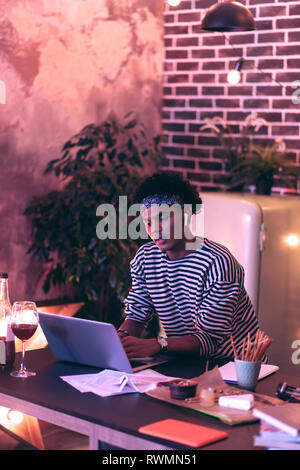 Teenager sitting at laptop and testing new headphones while pencils for notes standing near at the table. Stock Photo